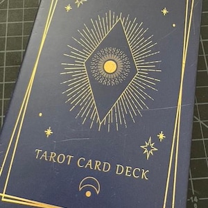May include: A navy blue tarot card deck box with gold foil accents. The box features a central diamond design with a sunburst pattern, stars, and the text "YOUR DREAM WILL COME TRUE" and "TAROT CARD DECK".