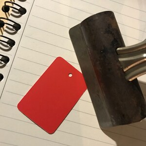 May include: A red rectangular tag with a hole in the center, lying on a white lined notebook next to a metal tool.