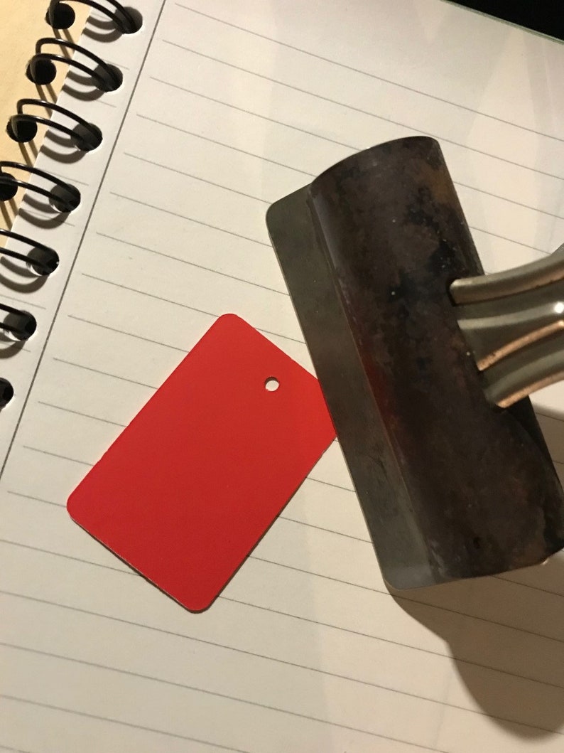 May include: A red rectangular tag with a hole in the center, lying on a white lined notebook next to a metal tool.