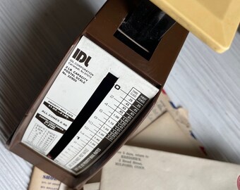 May include: A brown postal scale with a white dial and a black needle. The scale has a maximum capacity of 2 pounds and features a chart for calculating postage rates. The scale is sitting on top of a stack of envelopes.