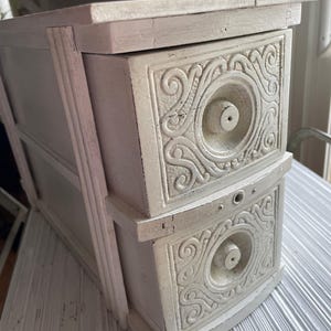 May include: White wooden two-drawer chest with ornate carved details on the front of each drawer. The chest has a distressed white paint finish.