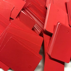 May include: A stack of bright red rectangular gift tags with a hole punched in the top corner. The tags are made of a sturdy cardstock and are ready to be used for gift wrapping.