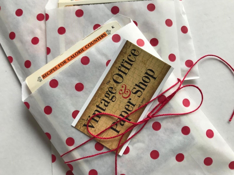 May include: A white paper bag with red polka dots is tied with a red string. The bag contains a stack of paper and a business card that reads "The Vintage Office & Paper Shop est. 2017".