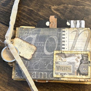 May include: A handmade journal with a cream-colored ribbon and tags. The journal features a vintage design with a dark gray background and large white numbers. A small rectangular image on the front reads "Writing" and depicts an ink bottle.