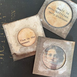 May include: Three square, rustic-style coasters. One coaster features a portrait, another reads "Live in the moment," and the third says "dwell in possibilities... emily dickinson." The coasters have a distressed, vintage aesthetic.