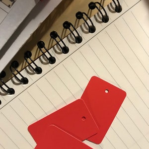 May include: Three red rectangular gift tags with a hole punched in the top corner. The tags are stacked on top of each other and are lying on a white lined notebook.