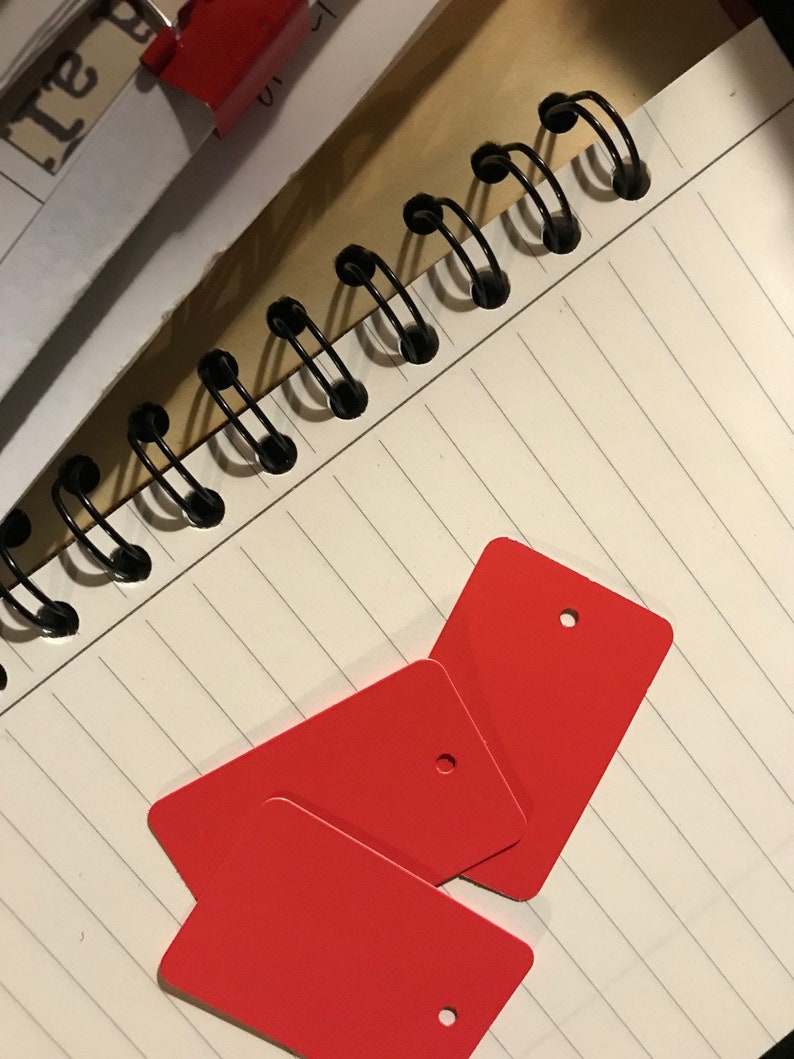May include: Three red rectangular gift tags with a hole punched in the top corner. The tags are stacked on top of each other and are lying on a white lined notebook.