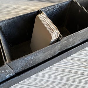 May include: A long, rectangular, metal container with multiple compartments. The container is dark gray with a weathered appearance. A stack of light-colored, rounded-corner cards sits in one compartment. The container is likely used for storage or organization.