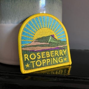 May include: An embroidered patch featuring a mountain scene with a rising sun behind it, in shades of yellow, green and blue. The text "ROSEBERRY TOPPING*" is embroidered in yellow on the patch.