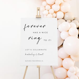 May include: A white sign on a wooden easel with the text "forever has a nice ring to it!" in black script. The sign also includes the names "kimberley & stuart" and the date "06.27.20XX | PALM SPRINGS, CA". A balloon arch in shades of white, peach, and beige is in the background.