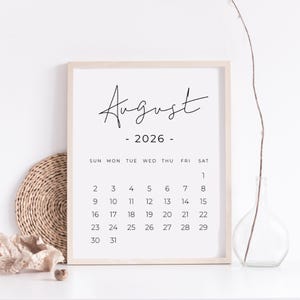 May include: A framed August 2026 calendar print. The calendar features a minimalist design with the month and year in elegant black script. The days of the week are listed across the top, with dates arranged below. The frame is a light wood color.