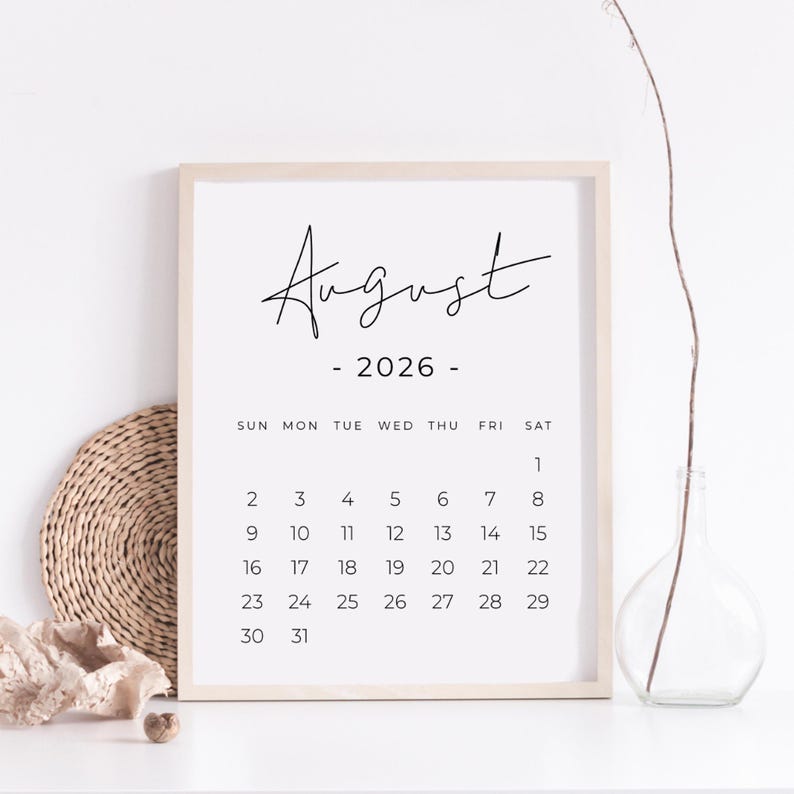 May include: A framed August 2026 calendar print. The calendar features a minimalist design with the month and year in elegant black script. The days of the week are listed across the top, with dates arranged below. The frame is a light wood color.