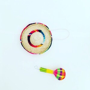 K&ouml;nnte beinhalten: Ein kleiner, bunter, gewebter Sombrero-Hut mit einer wei&szlig;en Schnur und eine bunte Maraca mit gelben, gr&uuml;nen, roten und blauen Streifen.