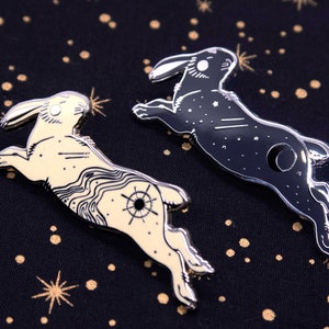 以下が含まれることがあります： 跳躍するスタイリッシュなウサギを描いたエナメルピン2個。1つのピンは白地に黒の星、もう1つのピンは黒地に白の星。両方のピンは黒のアウトラインです。
