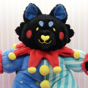 Puede incluir: Un peluche con forma de oso negro con orejas azules y detalles amarillos. El oso lleva un cuello de bufón rojo y amarillo y un atuendo a rayas azules y blancas. Tiene mejillas rojas y una nariz amarilla en forma de corazón.