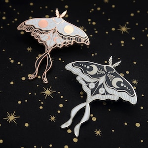 May include: Two moth-shaped enamel pins. One pin is light gray with rose gold accents and a copper-colored outline. The other pin is black and white with silver accents. Both pins have moon and star designs.