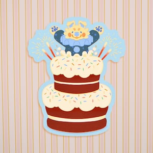 May include: A two-tiered birthday cake illustration with a light blue border. The cake is brown with white frosting and colorful sprinkles. A cartoon character with a blue outfit and yellow face is on top of the cake, with fireworks and candles.