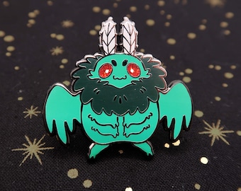 May include: A green and black enamel pin of a cartoon creature with large red eyes, feathery antennae, and wings. The creature has a furry green body and is standing on its hind legs.
