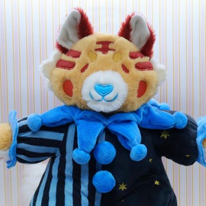 May include: A plush toy tiger in a clown costume. The tiger has a tan face with red markings, white fur, and a blue heart-shaped nose. It wears a blue and black striped outfit with blue pom-poms and stars.
