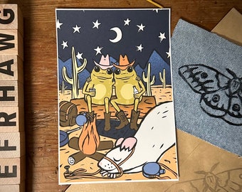 May include: A whimsical illustration of two frogs wearing cowboy hats sitting by a campfire under a starry night sky. A white rat with a pink nose is sleeping nearby. The illustration is printed on a white card.