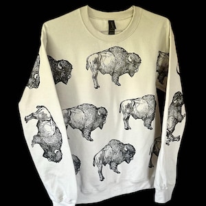 May include: A cream-colored sweatshirt featuring multiple black and white illustrations of bison. The bison are depicted in various poses, creating a repeating pattern across the garment. The sweatshirt has long sleeves and a crew neck.