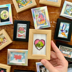 Pode incluir: Uma coleção de miniaturas emolduradas com várias ilustrações caprichosas, incluindo sapos, um porco, um unicórnio e um pássaro. As obras de arte são exibidas em uma superfície de madeira.