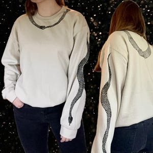 Op de afbeelding: Een wit sweatshirt met een zwart slangontwerp op de mouw. Het slangontwerp zit op beide mouwen.