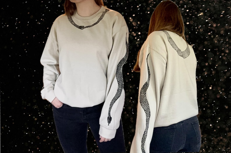 Op de afbeelding: Een wit sweatshirt met een zwart slangontwerp op de mouw. Het slangontwerp zit op beide mouwen.