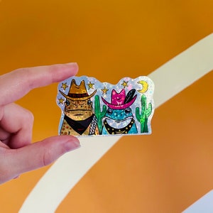 May include: A holographic sticker featuring two frogs wearing cowboy hats and bandanas. The frogs are standing in front of cacti and stars. The sticker has a glittery finish.