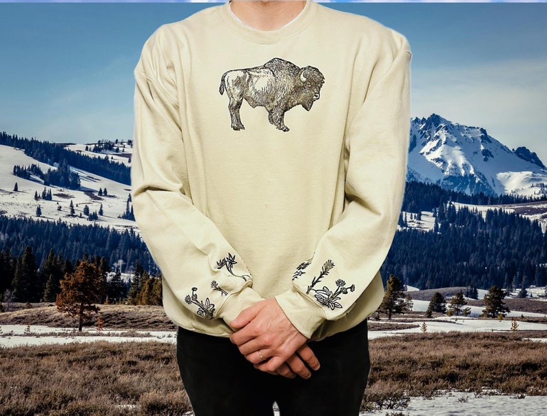 May include: A beige sweatshirt with a black and white illustration of a buffalo on the front. The sleeves have a black and white floral design.