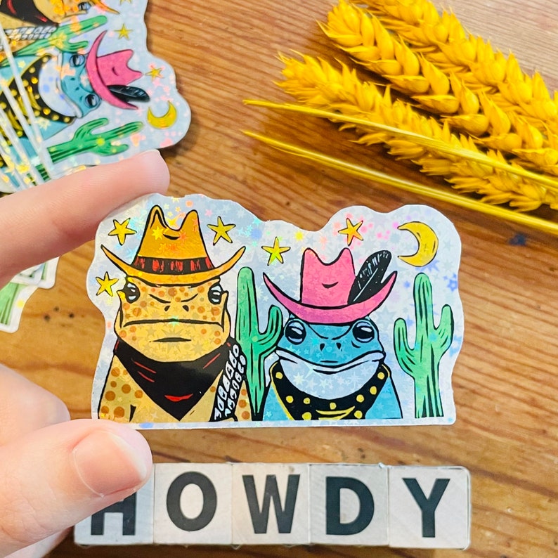 May include: Two frog stickers with a holographic finish. The frogs are wearing cowboy hats and bandanas. The sticker is set against a background of stars and a crescent moon. The text "HOWDY" is visible below the sticker.