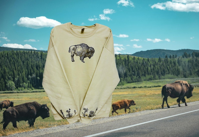 May include: A light beige sweatshirt with a black and white embroidered image of a buffalo on the front. The sleeves have embroidered floral designs on the cuffs. The sweatshirt is photographed against a backdrop of a grassy field, a road, and a forest.
