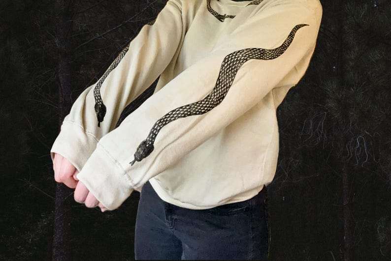 Op de afbeelding: Een cr&egrave;mekleurige trui met een zwart slangendesign op de mouwen. Het slangendesign is een herhalend patroon van schubben.
