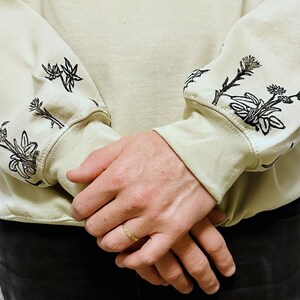 May include: A beige sweatshirt with black floral embroidery on the sleeves. The sweatshirt is folded and the sleeves are rolled up.