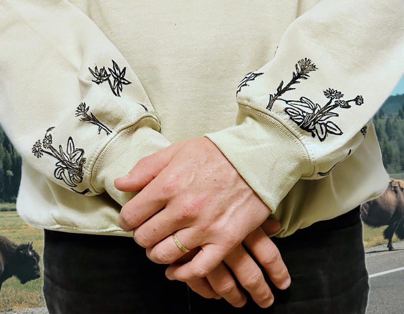 May include: A beige sweatshirt with black floral embroidery on the sleeves. The sweatshirt is folded and the sleeves are rolled up.