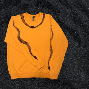 Op de afbeelding: Een feloranje longsleeve sweatshirt met een zwart slangendesign op de mouwen en de halslijn.
