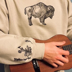 May include: A beige sweatshirt with a black and white illustration of a buffalo on the front. The sweatshirt has black and white floral embroidery on the sleeves. A person is holding a brown ukulele.
