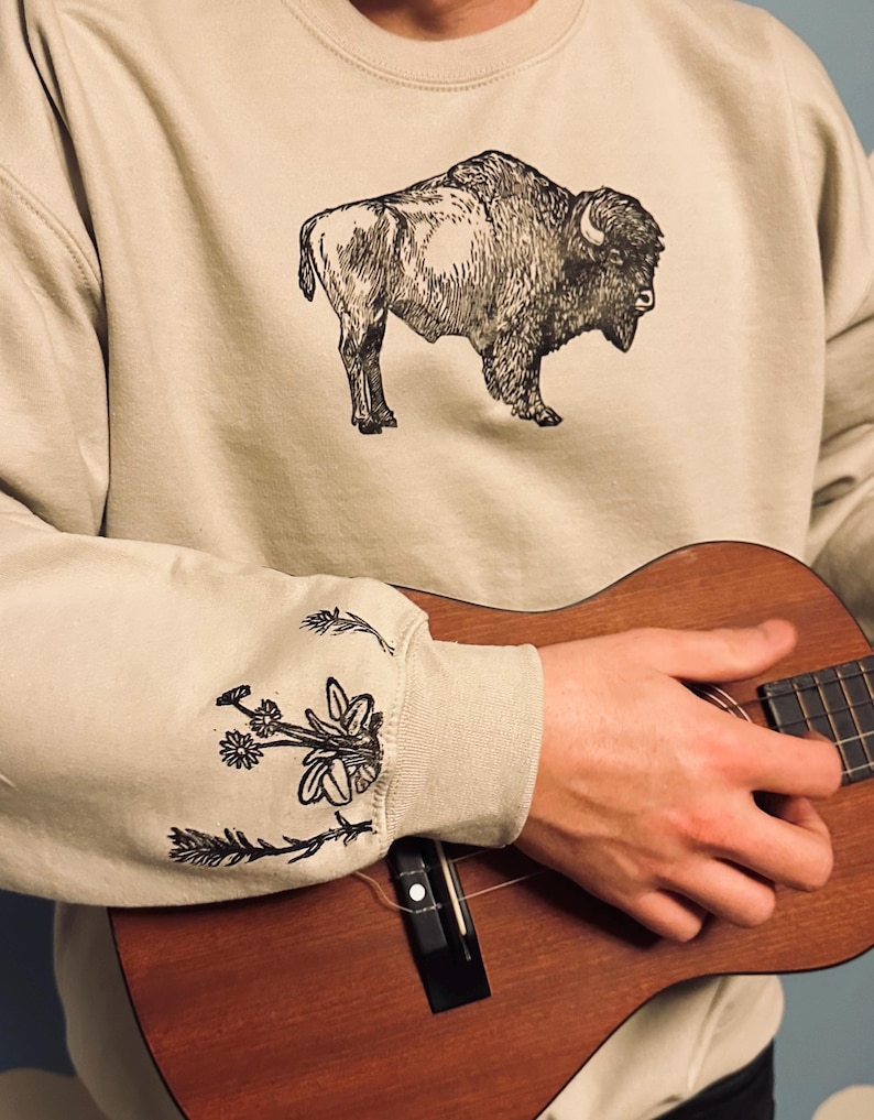 May include: A beige sweatshirt with a black and white illustration of a buffalo on the front. The sweatshirt has black and white floral embroidery on the sleeves. A person is holding a brown ukulele.