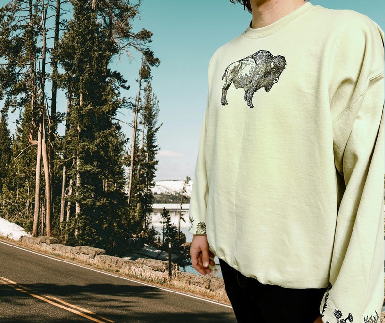 May include: A light yellow sweatshirt with a black and white graphic of a buffalo on the front. The sweatshirt has long sleeves and a crew neck.