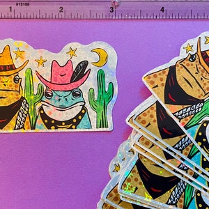 May include: A set of five iridescent holographic stickers featuring two frogs wearing cowboy hats and bandanas. The frogs are surrounded by cacti and stars. The stickers are perfect for adding a touch of whimsy to laptops, water bottles, or journals.