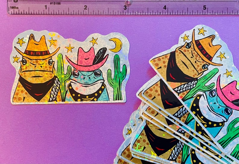 May include: A set of five iridescent holographic stickers featuring two frogs wearing cowboy hats and bandanas. The frogs are surrounded by cacti and stars. The stickers are perfect for adding a touch of whimsy to laptops, water bottles, or journals.