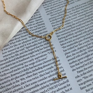 以下が含まれることがあります： T字型の留め具が付いたゴールドチェーンネックレス。チェーンは小さな正方形のリンクでできています。ネックレスは、本が置かれた白い背景に撮影されています。