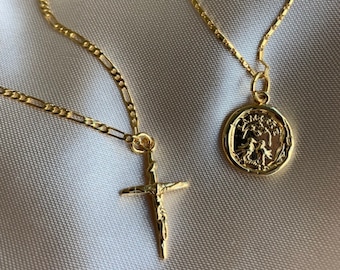 May include: Two gold-colored necklaces with pendants. One necklace features a crucifix pendant, and the other has a round pendant with a detailed design. The necklaces are displayed on a white, silky surface.
