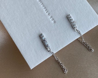 Puede incluir: Un par de pendientes de plata con una pequeña cadena que cuelga de cada uno. Cada pendiente tiene una barra con cinco pequeñas piedras transparentes.
