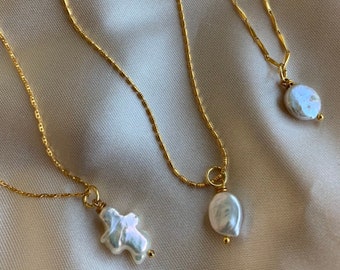 May include: Three gold necklaces with pearl pendants. The pearls are different shapes and sizes. The necklaces are on a beige fabric background.