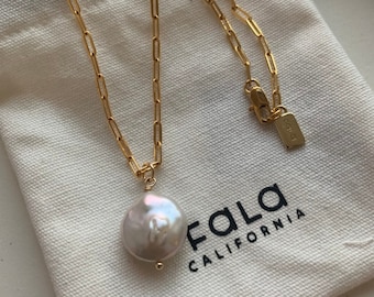 May include: A gold chain necklace with a large, white pearl pendant. The necklace is on a white fabric pouch with the text "FALA CALIFORNIA" printed on it.