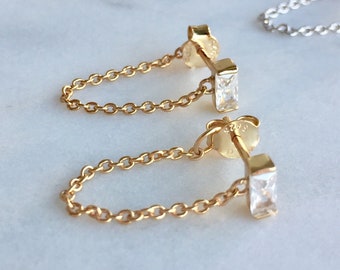 Puede incluir: Un par de pendientes de oro con una pequeña piedra transparente de forma cuadrada y una delicada cadena de oro que cuelga de cada pendiente.