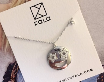 May include: Silver necklace with a round pendant featuring a smiley face design with star-shaped eyes. The pendant is embellished with clear crystals. The necklace is displayed on a white card with the brand name FALA and the website address SHOP.PLAYWITHFALA.COM.