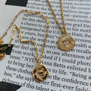 May include: Two gold necklaces with pendants. One pendant features a crescent moon with a star and small diamonds. The other pendant features a sun with an eye and stars. The necklaces are on a book with text about a Parsons grad.