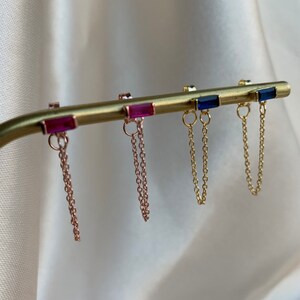 Könnte beinhalten: Drei Paar goldene Ohrringe mit einer baumelnden Kette. Die Ohrringe haben einen kleinen rechteckigen Stein in Pink, Blau und Blau.